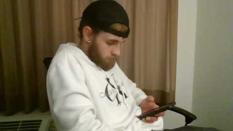 Media: A video of a bearded man in a white hoodie, black backward baseball cap, and hoop earrings, engrossed in a smartphone, seated in a beige room with curtains.