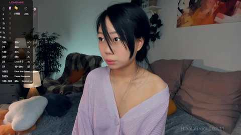 Media: A video of an East Asian woman with black hair in a messy bun, wearing a light purple cardigan, sitting on a bed in a dimly lit room.