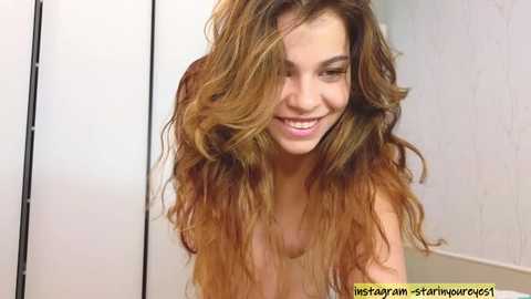 Media: Video of a young woman with long, wavy, light brown hair, smiling, wearing a sleeveless top, standing in front of a white wall and a partially visible mirror.