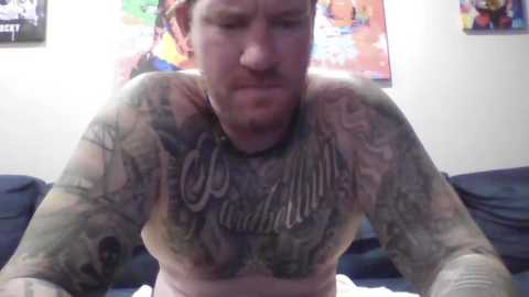 Media: Video of a shirtless man with extensive tattoos covering his arms and chest, wearing a choker necklace. Background includes colorful posters and a dark couch.