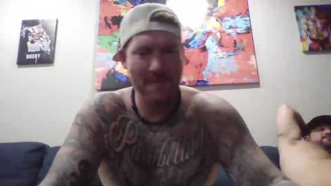 Media: Video of a tattooed man in a white t-shirt and backwards cap, grinning widely, with colorful abstract art and another person partially visible in the background.