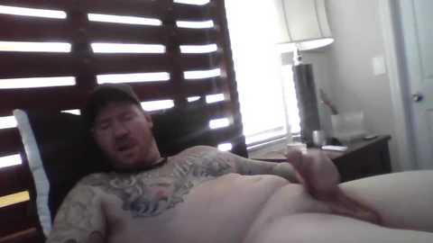 Media: Video of a tattooed man lying on a bed, masturbating, with a window and blinds in the background.