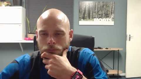 Media: Video of a bald, bearded man in a blue and black shirt, deep in thought, sitting at a desk with a black chair, gray wall with a \"NOMA\" sign, and a potted plant.