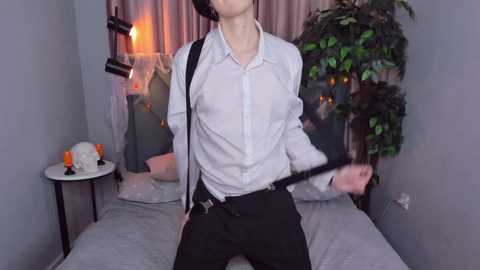 Media: Video of a slender, pale-skinned man in a white dress shirt, black pants, and black suspenders, striking a provocative pose with a whip in a dimly lit bedroom with pink curtains, a lamp, and a potted plant.