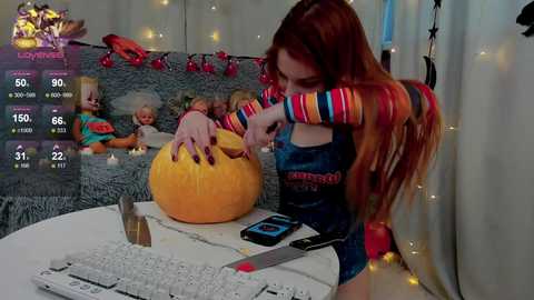 Media: Video of a red-haired woman in a blue jumpsuit, carving a pumpkin on a table. Background features a plush toy and a keyboard.