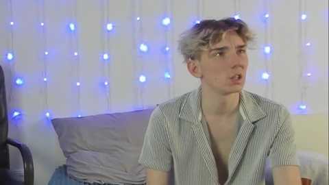 Media: Video of a pale, curly-haired young man in a striped shirt, looking surprised, against a backdrop of glowing blue fairy lights and a white wall.