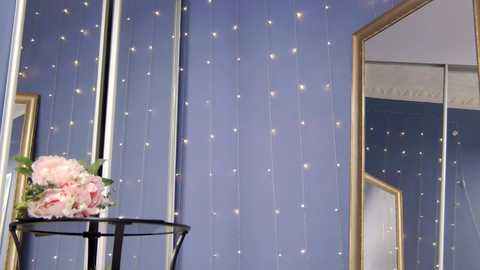 Media: Video of a room with a purple wall adorned with vertical gold stripes and sparkling fairy lights, featuring a black metal stand holding pink peonies and a gold-framed mirror.