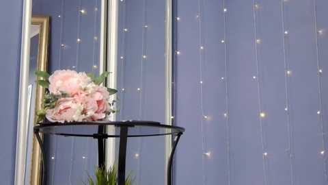 Media: Video of a modern interior with a lavender wall adorned with string lights. A black metal stand holds a bouquet of pink peonies and greenery, with a mirror reflecting part of the room.