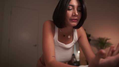 Media: Video of a young Asian woman with straight black hair, wearing a white tank top, sitting in a dimly lit room, focusing on a laptop.