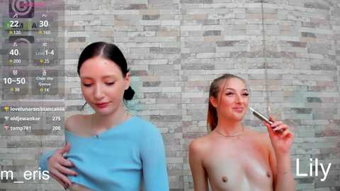 Media: Video of two young women in a bathroom. One, with fair skin and dark hair, wears a blue top; the other, with fair skin and blonde hair, is topless, holding a lipstick.