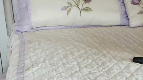 Media: A video of a neatly made bed with a white quilted bedspread, lavender pillowcases, and a floral-patterned pillow, featuring a soft purple rose. The background shows a plain white wall.