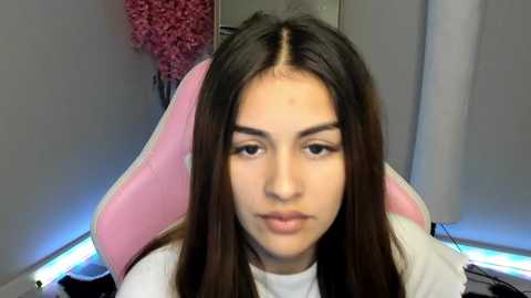 Media: Video of a young woman with long brown hair, fair skin, and a neutral expression, seated in a pink gaming chair. Background features a light-colored wall, pink artificial flowers, and a blue LED strip.