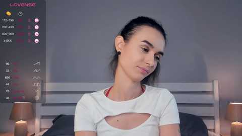 Media: A video of a young woman with light skin, brown hair tied back, wearing a white crop top and sitting on a bed. The background shows a gray wall and bedside lamps.