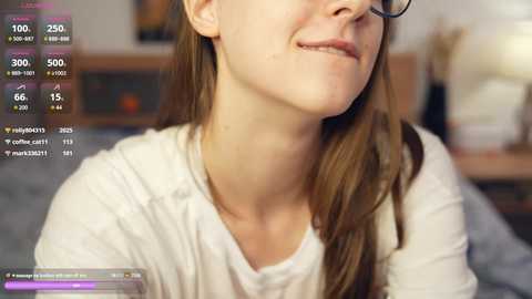 Media: Video of a fair-skinned woman with glasses, long brown hair, and a slight smile. She wears a white long-sleeved shirt. Background shows a blurred living room with a TV and books.