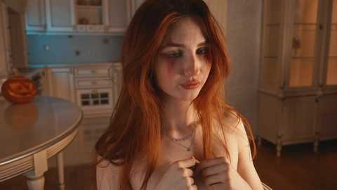 Media: A video of a young woman with long, auburn hair, pale skin, and rosy cheeks, wearing a white top, seated in a softly lit, vintage kitchen with light blue cabinets and wooden floors.