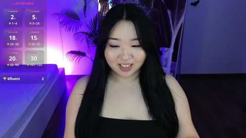 Media: Video of a smiling Asian woman with long black hair, wearing a black strapless top, indoors with purple lighting, and a digital clock showing 18:18.