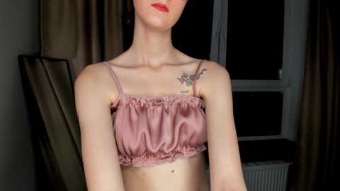 Media: Video of a slender, fair-skinned woman with dark hair wearing a pink satin crop top, showcasing a delicate flower tattoo on her left shoulder. Background features a window with dark curtains and a radiator.