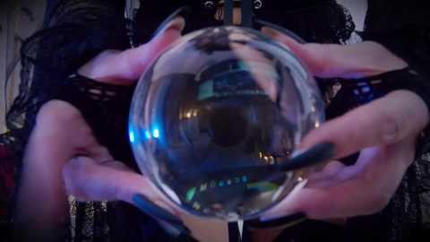 Media: Video of a person's hands holding a clear, reflective orb, showcasing their dark, intricate lace gloves and a hint of their black dress. The background is blurred, focusing attention on the hands and orb.