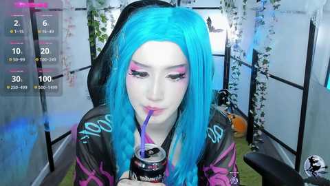 Media: A video of a young woman with vibrant blue hair, wearing a black hoodie with pink accents, drinking from a can, in a dimly lit, modern room with white walls and plants.