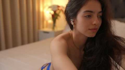 Media: Video of a young woman with long, dark hair and light skin, wearing a strapless blue and white dress, lying on a bed in a softly lit bedroom with beige curtains and a nightstand.