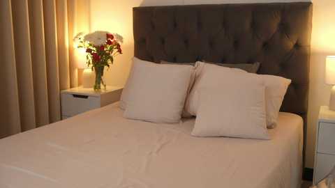 Media: Video of a neatly made bed with beige sheets, two pillows, and a tufted dark headboard. A white nightstand with a lamp and vase of red and white flowers is on the left side.