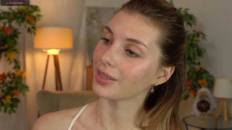 Media: Video of a fair-skinned woman with light brown hair, wearing a white top, smiling softly, standing in a warmly lit living room with a beige couch, a lamp, and floral decorations.