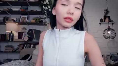 Media: Video of an Asian woman with long black hair, fair skin, wearing a sleeveless light blue top, standing in a bookshelf-filled, cozy bedroom with a hanging lamp and plants.