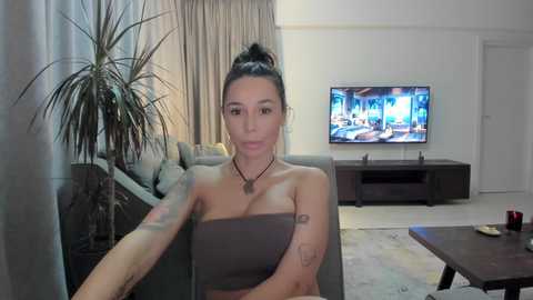 Media: Video of a woman with medium skin tone and dark hair in a bun, wearing a strapless grey top, sitting on a grey couch in a modern living room with a flat-screen TV and potted plant.