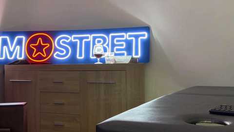 Media: Video of a minimalist bedroom with a large, glowing neon \"MOSTREET\" sign, a wooden dresser, a bed with a gray sheet, and a bedside table with a lamp and remote control.