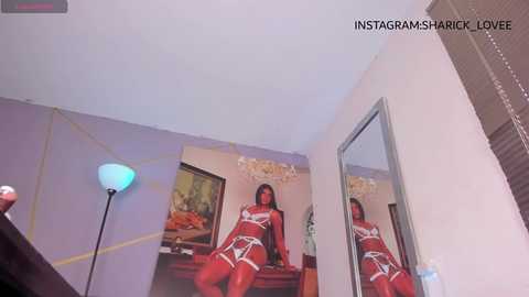 Media: Video of a woman in red lingerie with white accents, sitting on a bed in a dimly lit room with a chandelier and a blue floor lamp.