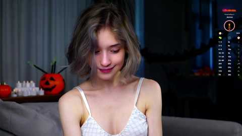 Media: Video of a young woman with short, messy blonde hair, wearing a white lace bra, sitting indoors, surrounded by Halloween decorations including a pumpkin and a calendar.