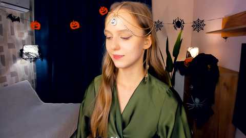 Media: Video of a young woman with long brown hair wearing a green robe and a gold crown, standing in a Halloween-themed room with dark walls, spider decorations, and a black coat.