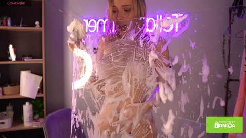 Media: Video of a blonde woman with medium breasts, covered in white body paint, standing in front of a neon sign in a dimly lit room.