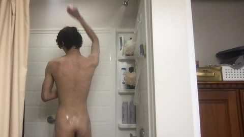 Media: Video of a naked, fair-skinned man with curly brown hair, mid-shower, facing away, in a white-tiled bathroom with a wooden cabinet and beige towels.