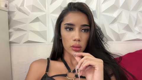 Media: Video of a young woman with long, dark hair, wearing a black leather harness top. She has a medium complexion, full lips, and is holding a silver key. Background features a white, geometric-patterned wall.