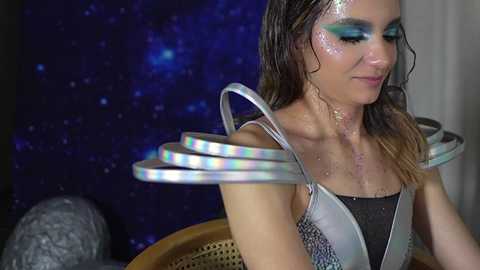 Media: A video of a woman with wet, shoulder-length brown hair wearing a silver sequined top, metallic shoulder guards, and glittery makeup. The background features a dark blue, starry night sky.