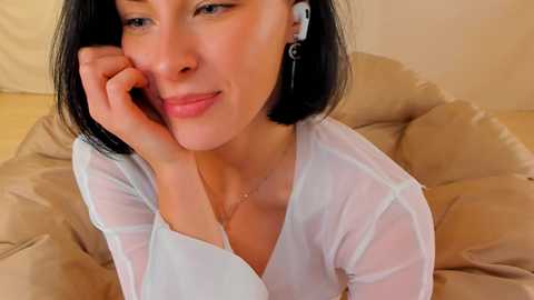 Media: Video of a smiling, fair-skinned woman with short black hair, wearing a white sheer top and silver earrings, lying on a beige cushioned chair, earbuds in, looking relaxed and content.