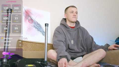 Media: Video of a young man with short hair, wearing a grey hoodie and beige shorts, meditating on a beige couch with a cardboard box. A digital overlay shows fitness metrics.