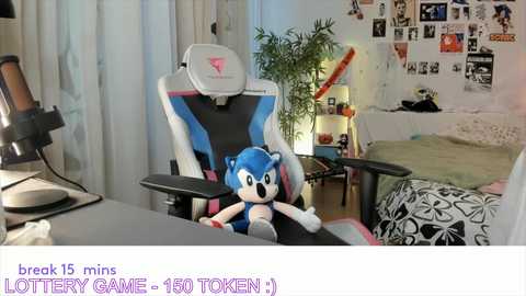 Media: Video of a Sonic the Hedgehog plush doll sitting in a white and blue gaming chair with a pink and black desk, surrounded by a cluttered room with posters and a plant.