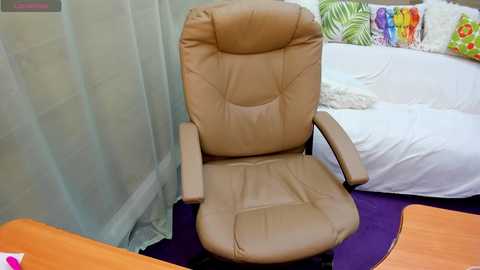 Media: Video of a tan leather executive chair with armrests, positioned on a purple carpet in a room with white curtains and a white sofa adorned with colorful pillows.