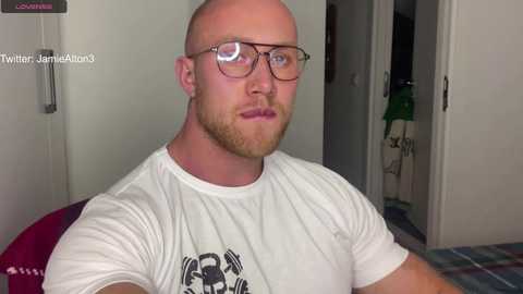 Media: Video of a muscular, bald, bearded man with glasses, wearing a white T-shirt with a black design, standing indoors.