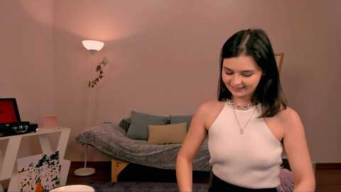 Media: Video of a woman with straight black hair, wearing a white sleeveless top and black pants, standing in a cozy, dimly lit living room with a gray couch, laptop, and lamp.