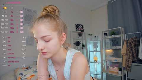 Media: Video of a young, fair-skinned woman with blonde hair in a bun, wearing a white tank top, in a cozy, cluttered bedroom with shelves and a bed.