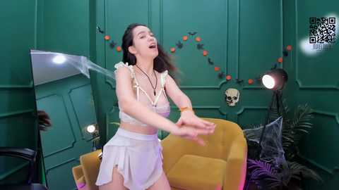 Media: Video of a young Asian woman with long black hair, wearing a white, frilly dress, dancing energetically in a green-paneled room decorated with Halloween elements.