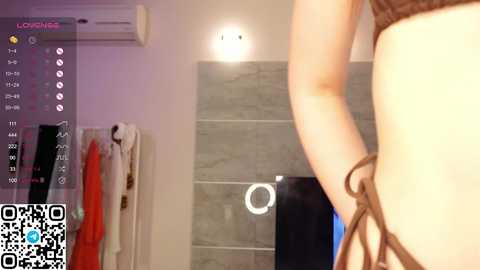 Media: A video of a woman in a brown bra, taken from a low angle, in a modern bathroom with a shower and gray tiles.