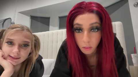 Media: Video of two young women with fair skin and red hair in a modern, minimalistic bathroom. The woman on the right has long, straight hair, dark makeup, and a black shirt, while the woman on the left has pigtails and a neutral expression.