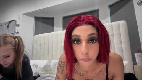 Media: Video of a woman with bright red hair, wearing black lingerie, tattoos on her arms, sitting on a bed with a white headboard, partially obscured by another person in the background.