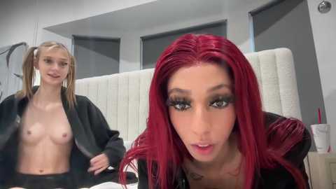 Media: Video of two women in a modern bathroom: One topless with small breasts, wearing a black jacket, blonde hair in pigtails. The other, with red hair, wearing a black jacket, has a shocked expression.