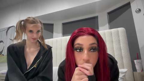 Media: Video of two young women in a modern, minimalist room. One has long red hair, wearing a black hoodie, and holds her finger to her lips. The other has blonde pigtails, a black jacket, and is looking at the camera.