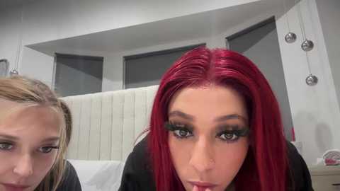 Media: Video of two women with long, straight red hair, wearing dramatic black eyeliner and mascara, seated in a modern, white-walled room with a bed in the background.